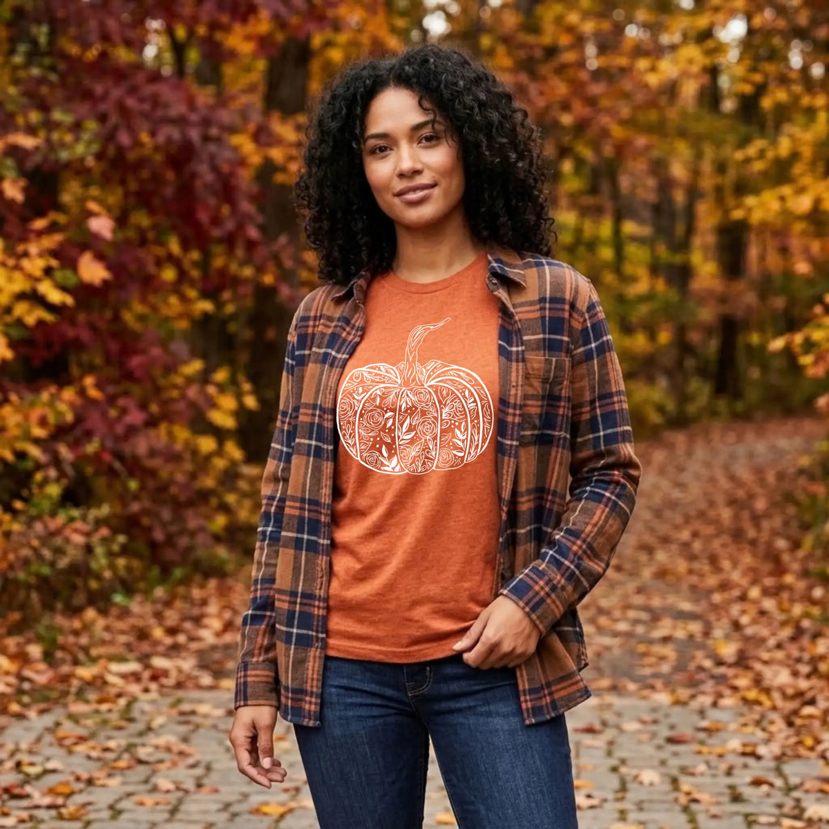 Woman in a heather autumn orange floral pumpkin graphic tee layered under a plaid flannel on a leafy fall path