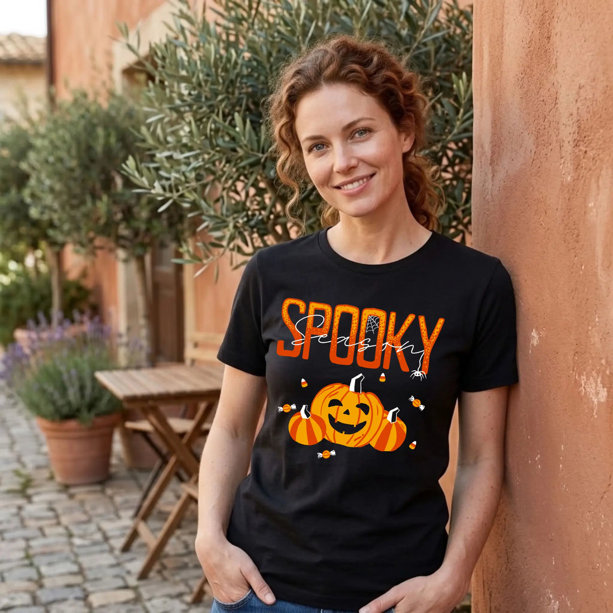 Smiling woman in a black "Spooky Season" Halloween graphic tee leaning against a terracotta wall