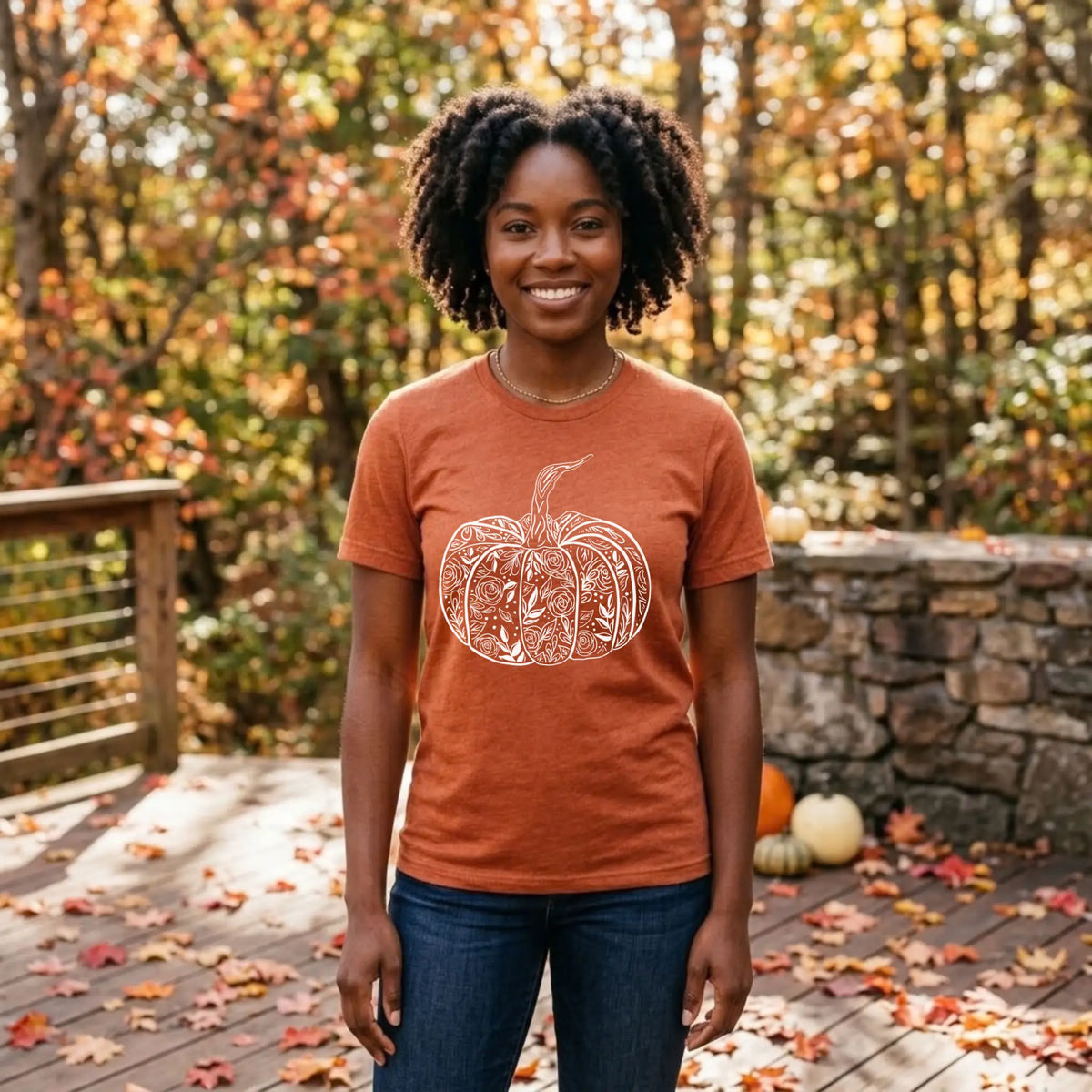 Smiling woman in a heather autumn orange floral pumpkin graphic tee on a wooden deck with fall leaves