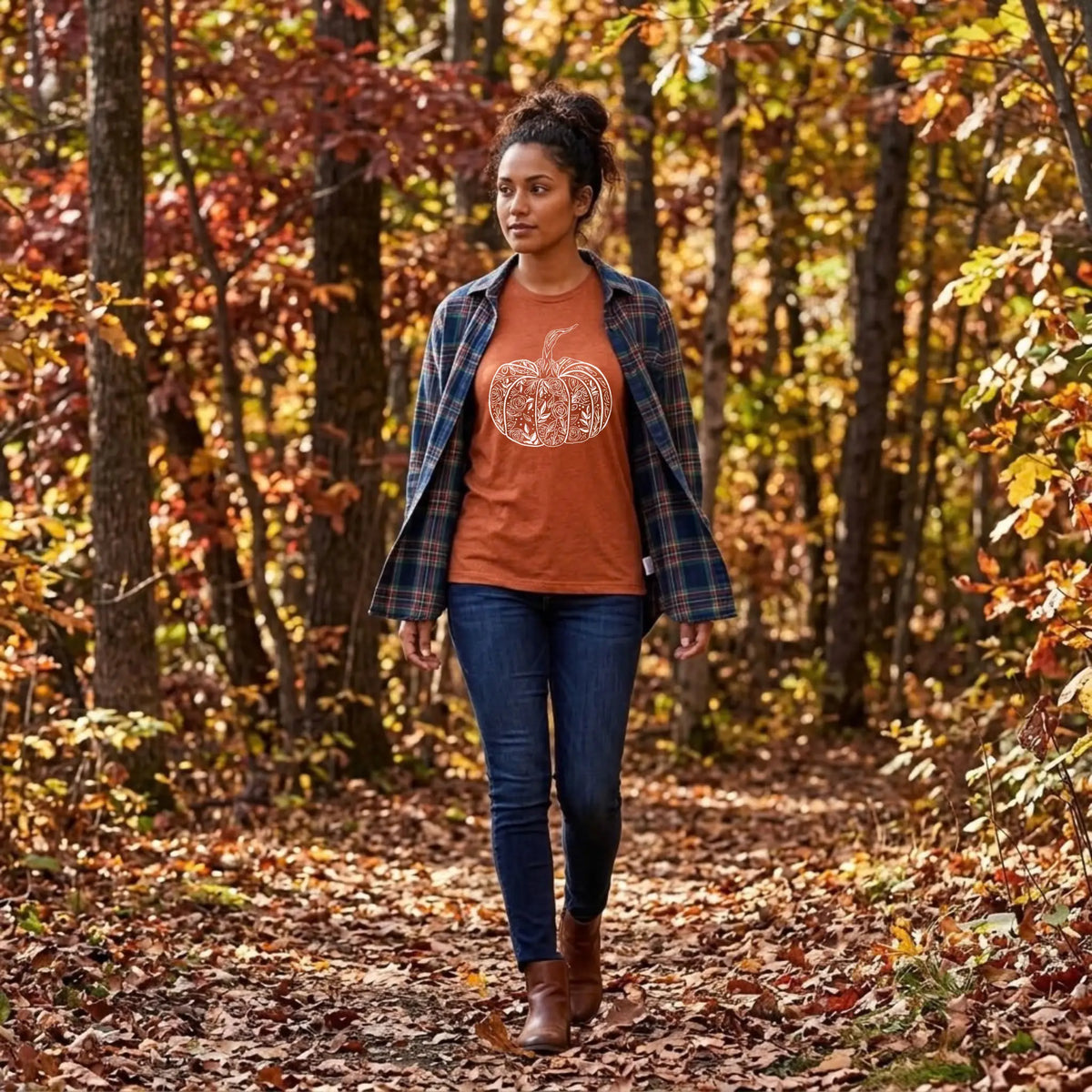 Woman in a heather autumn orange floral pumpkin graphic tee and navy plaid flannel walking a wooded fall trail