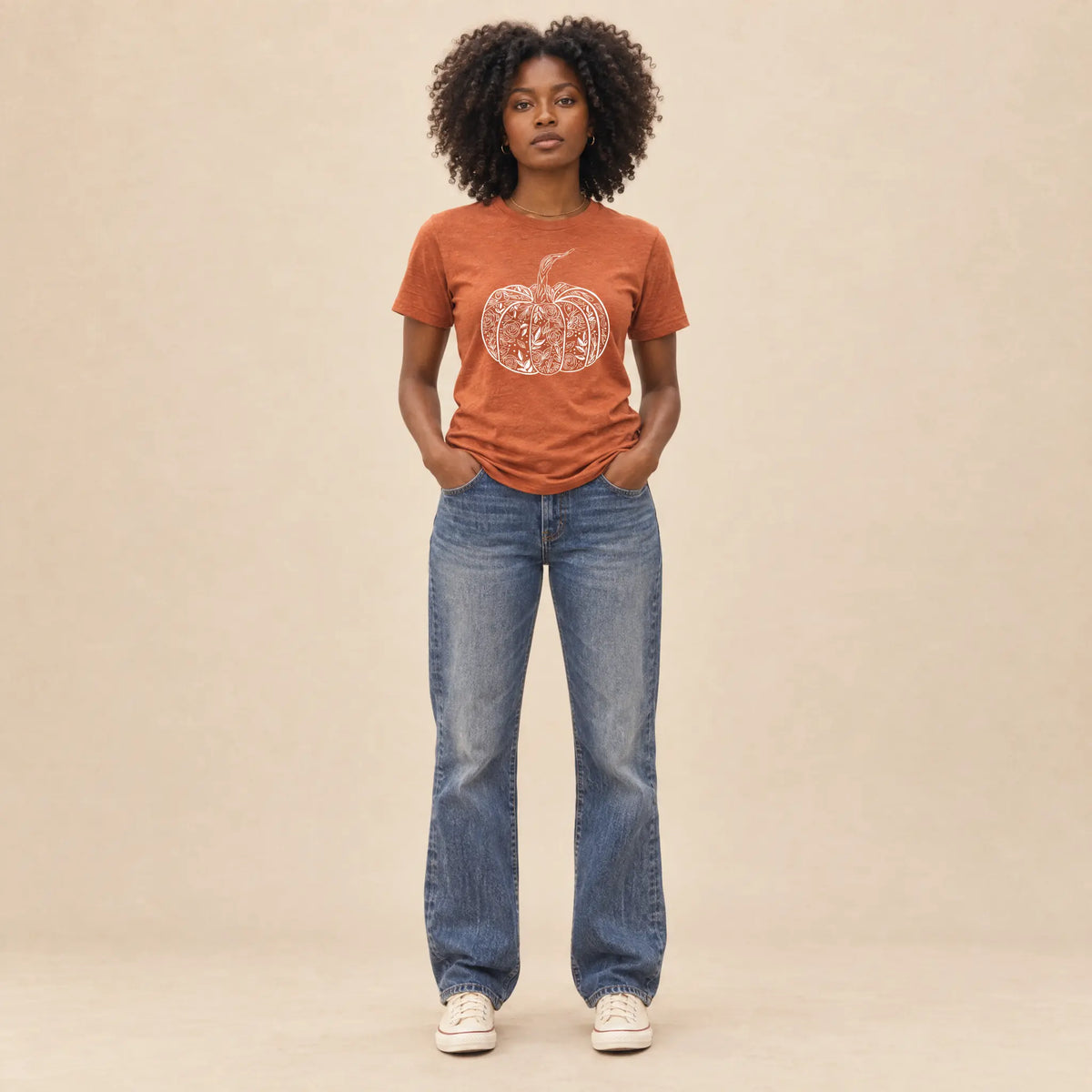 Full-length view of a woman in a heather autumn orange floral pumpkin graphic tee and straight leg jeans on a neutral backdrop
