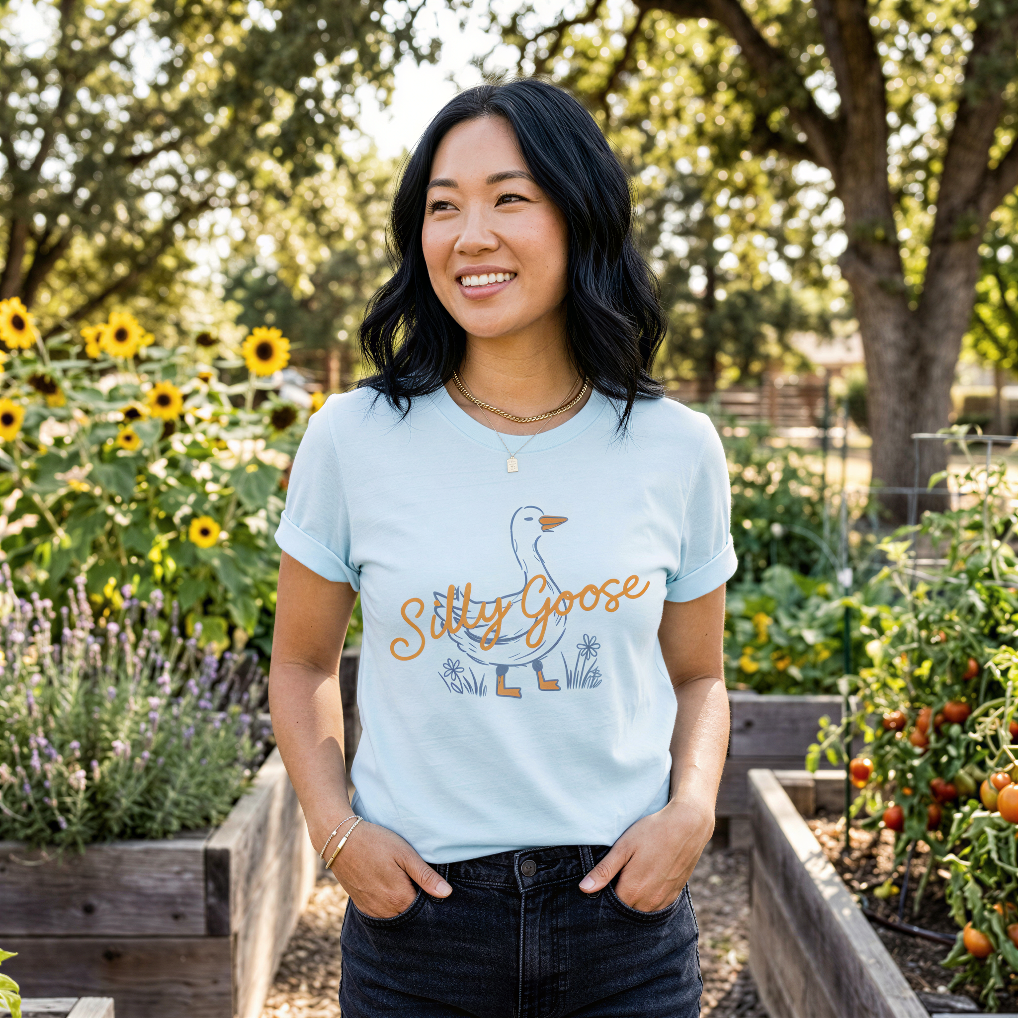 A woman wearing a light blue t-shirt with a graphic of a goose and the text "Silly Goose" stands in a garden surrounded by flowers and plants.