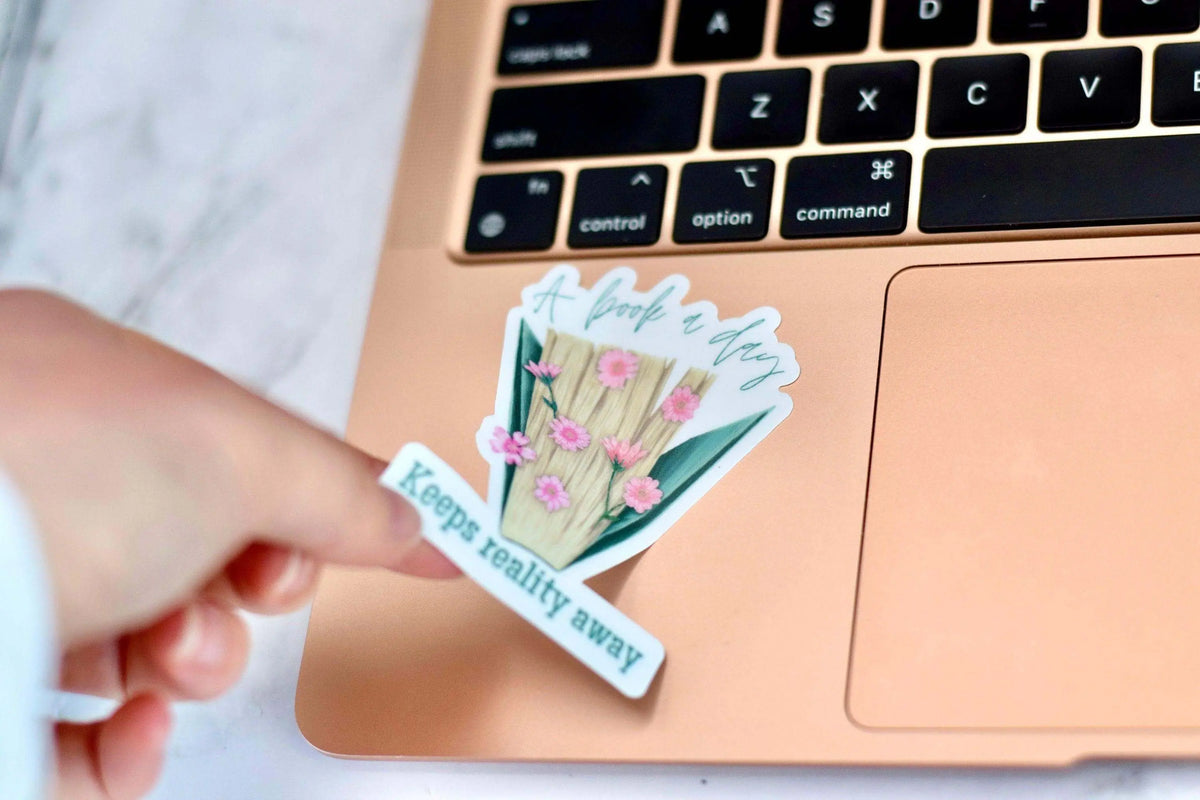 Vinyl sticker featuring the quote "A Book a Day Keeps Reality Away" by Winks Design Studio, LLC, with colorful book and whimsical design.