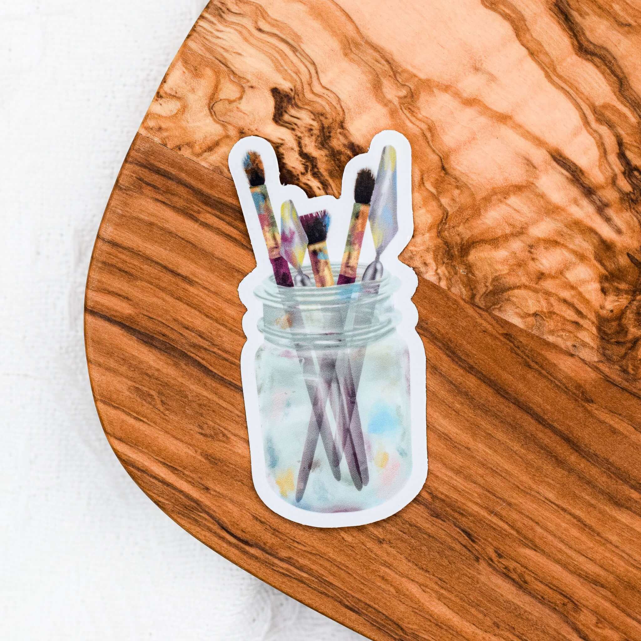 Vinyl sticker of paint brushes in a glass jar by Winks Design Studio, LLC, featuring realistic brush details on a clear background.