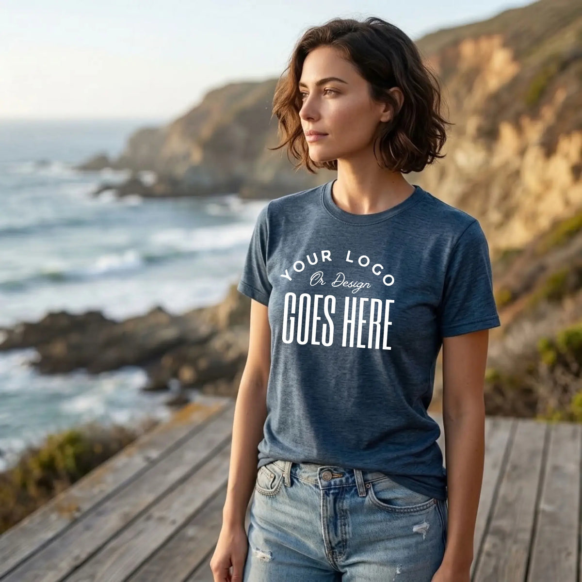 Woman in a heather navy custom t-shirt with a large white "Your Logo or Design Goes Here" center chest placeholder on a coastal overlook