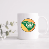 SCBA Ceramic Mug with custom logo on a light background àdorned with greenery