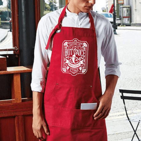 Man wearing red NC Hot Sauce Contest & Festival apron with vintage label design, ideal for cooking and grilling.