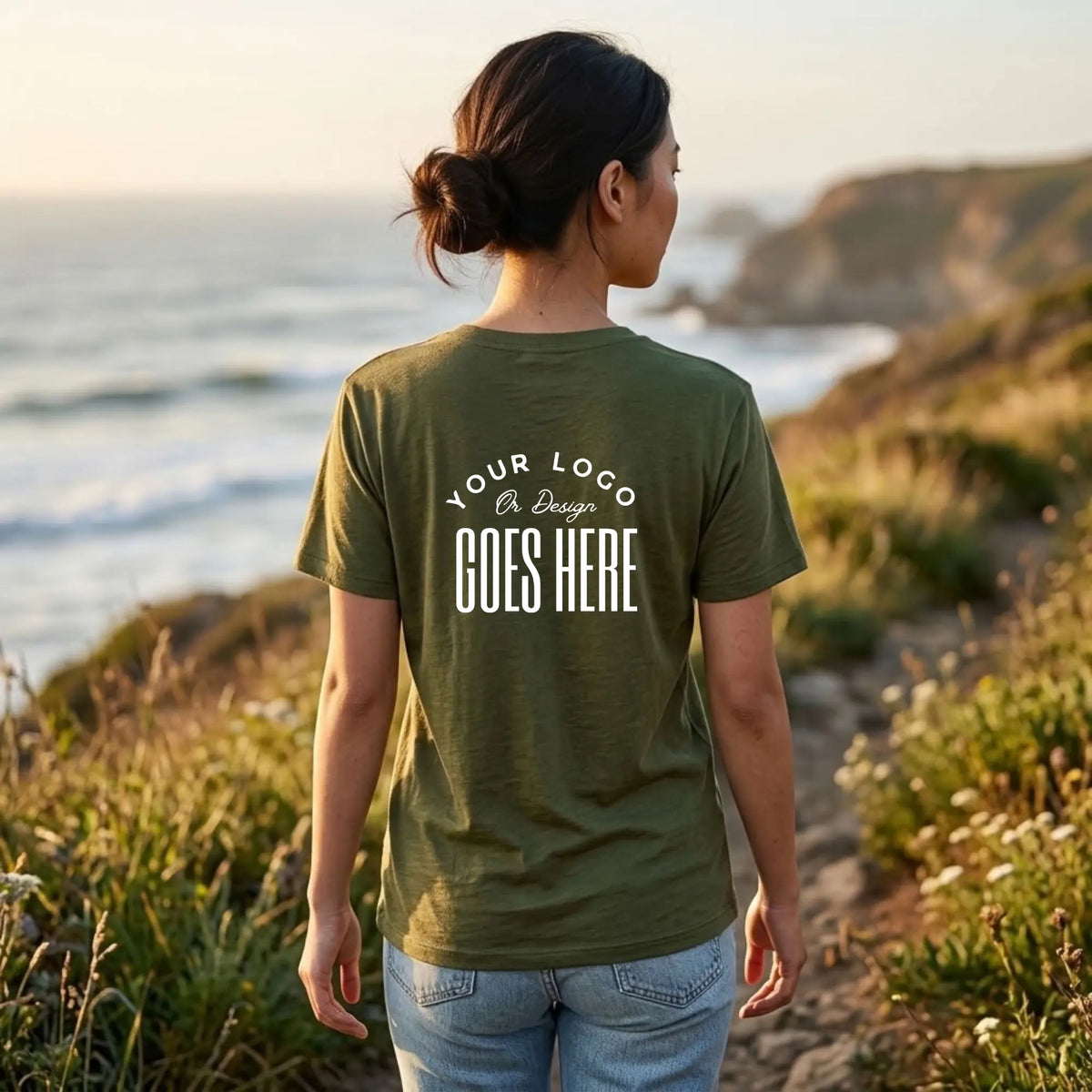 Back view of a woman in a military green custom t-shirt with a large white "Your Logo or Design Goes Here" back print on a coastal trail