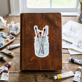Brown leather notebook with a mason jar illustration of paintbrushes on a wooden surface with art supplies.