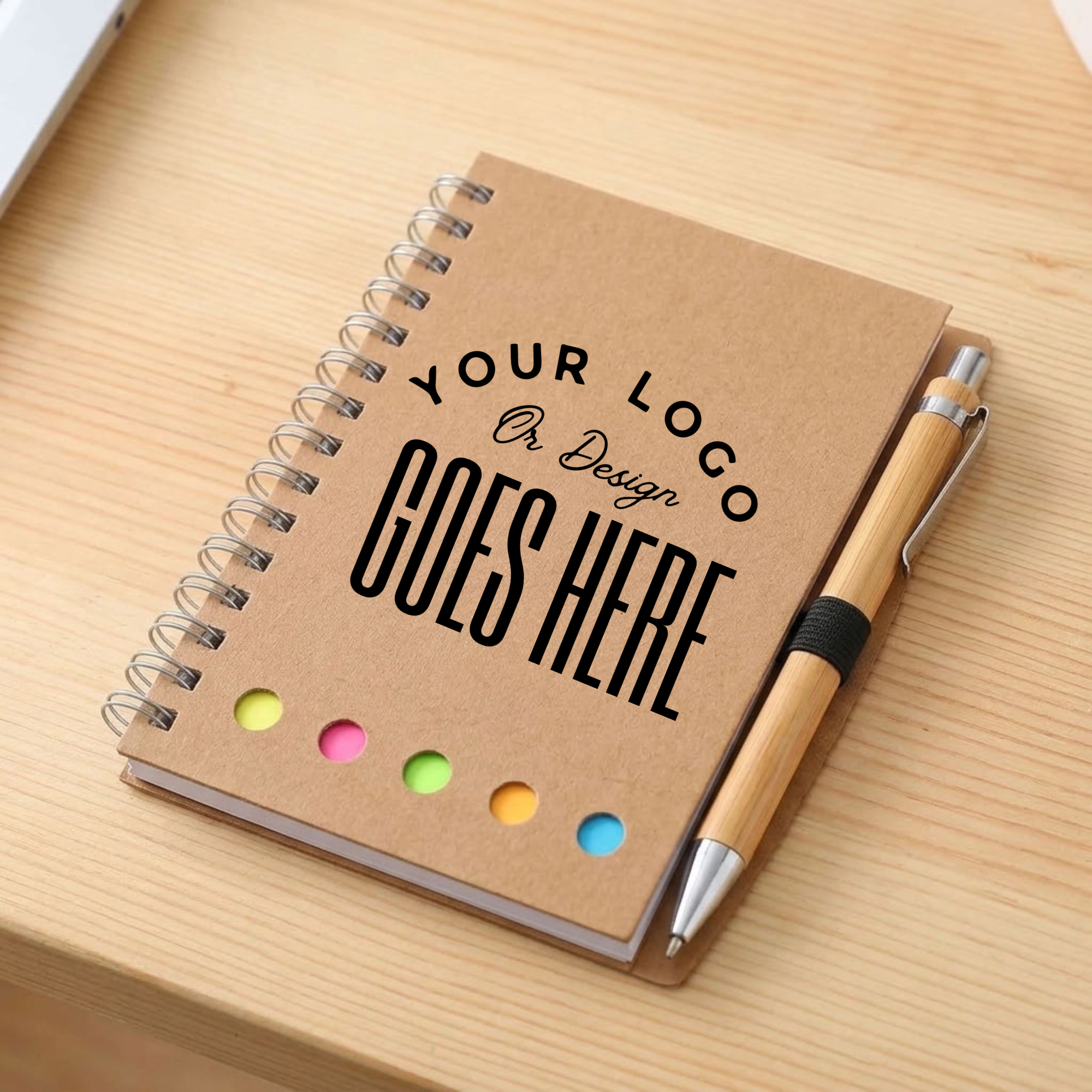 A spiral-bound kraft paper notebook on a light wood desk, featuring customizable black text and five colorful dot accents. A bamboo pen with a black band lies beside it.