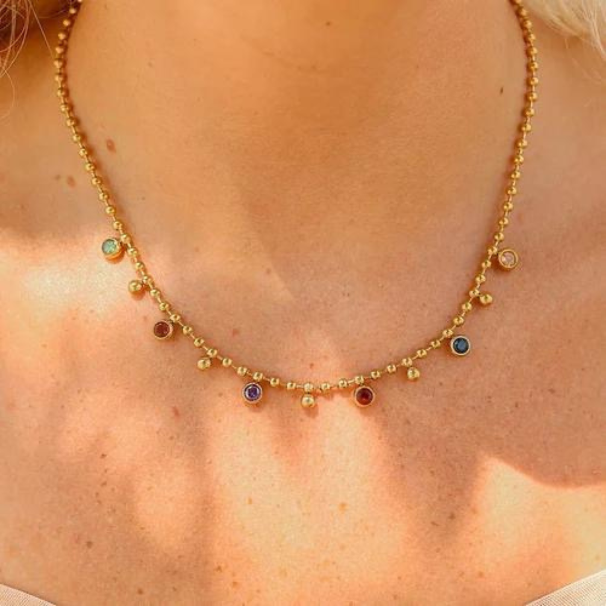 A close-up of a gold beaded necklace with small colorful gemstone accents worn on a model&