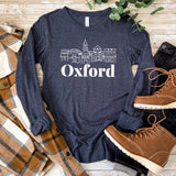 Navy long sleeve shirt with Oxford skyline design, paired with plaid shirt and brown boots, laid out on wooden surface.