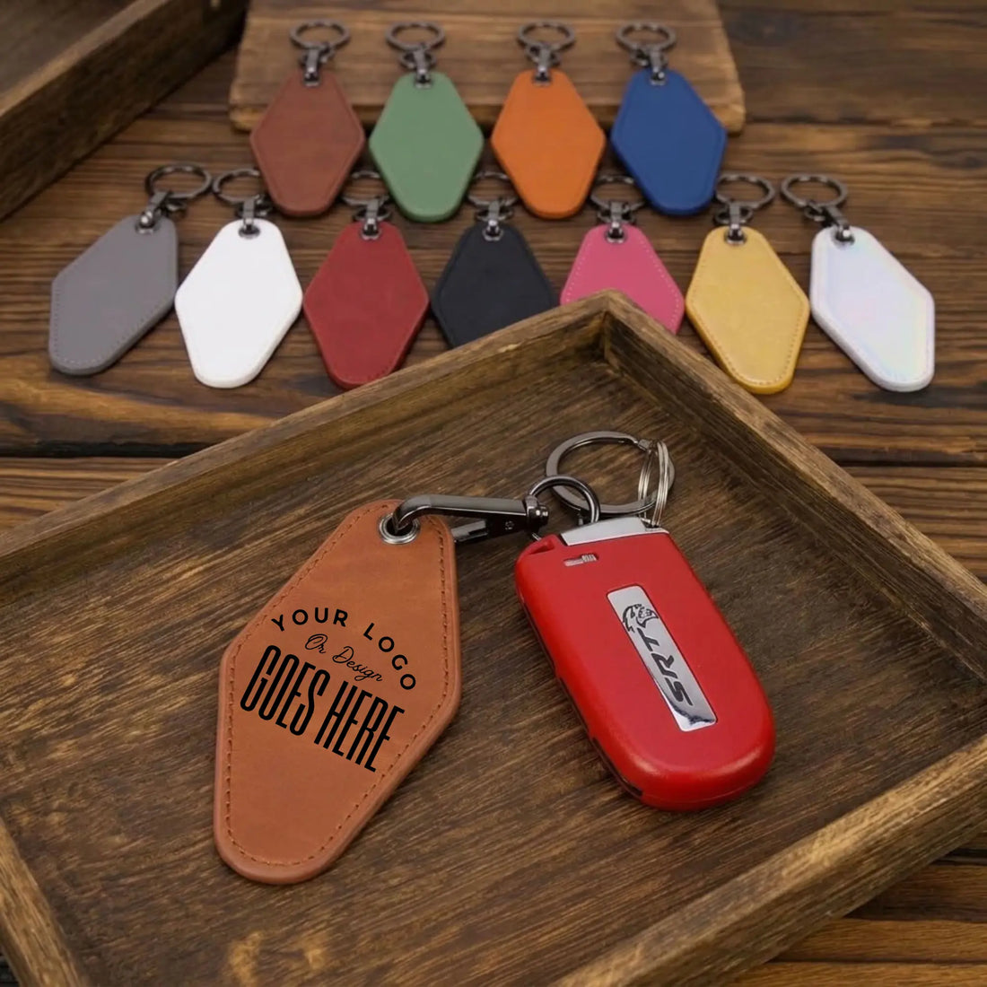 A set of customizable leather keychains displayed on a dark wooden tray, with one brown leather keychain in the foreground showing placeholder text and a red car key fob next to it. Additional color options are lined up in the background.