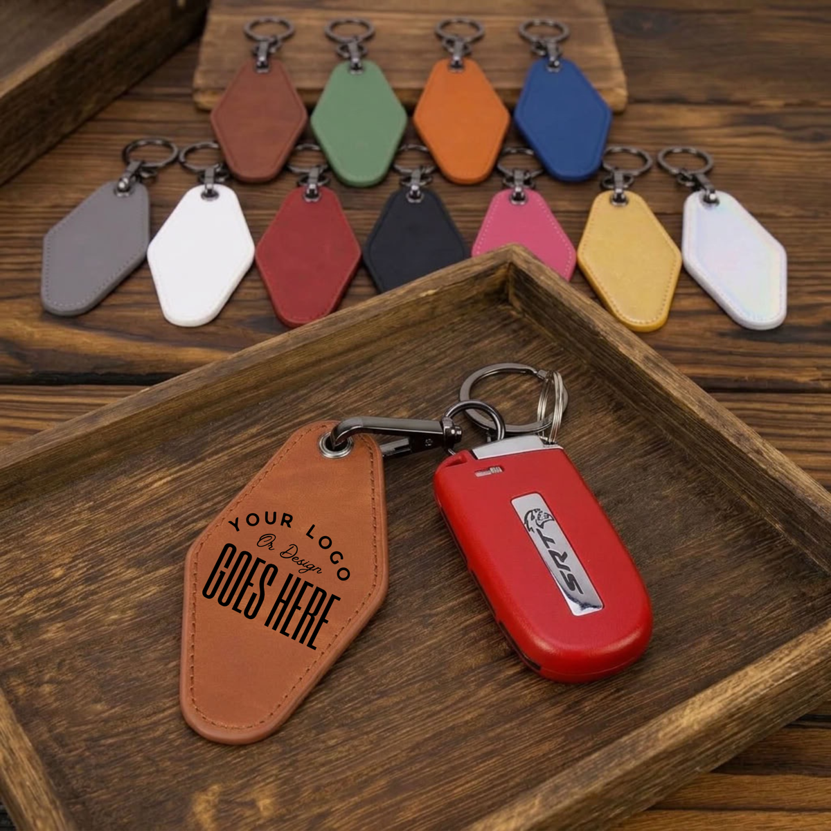 A set of customizable leather keychains displayed on a dark wooden tray, with one brown leather keychain in the foreground showing placeholder text and a red car key fob next to it. Additional color options are lined up in the background.