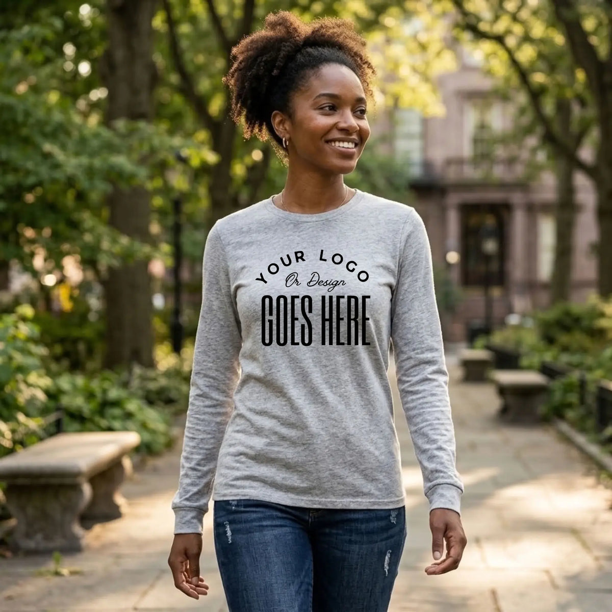 Woman in a heather gray custom long sleeve t-shirt with a large black "Your Logo or Design Goes Here" center chest placeholder in a park