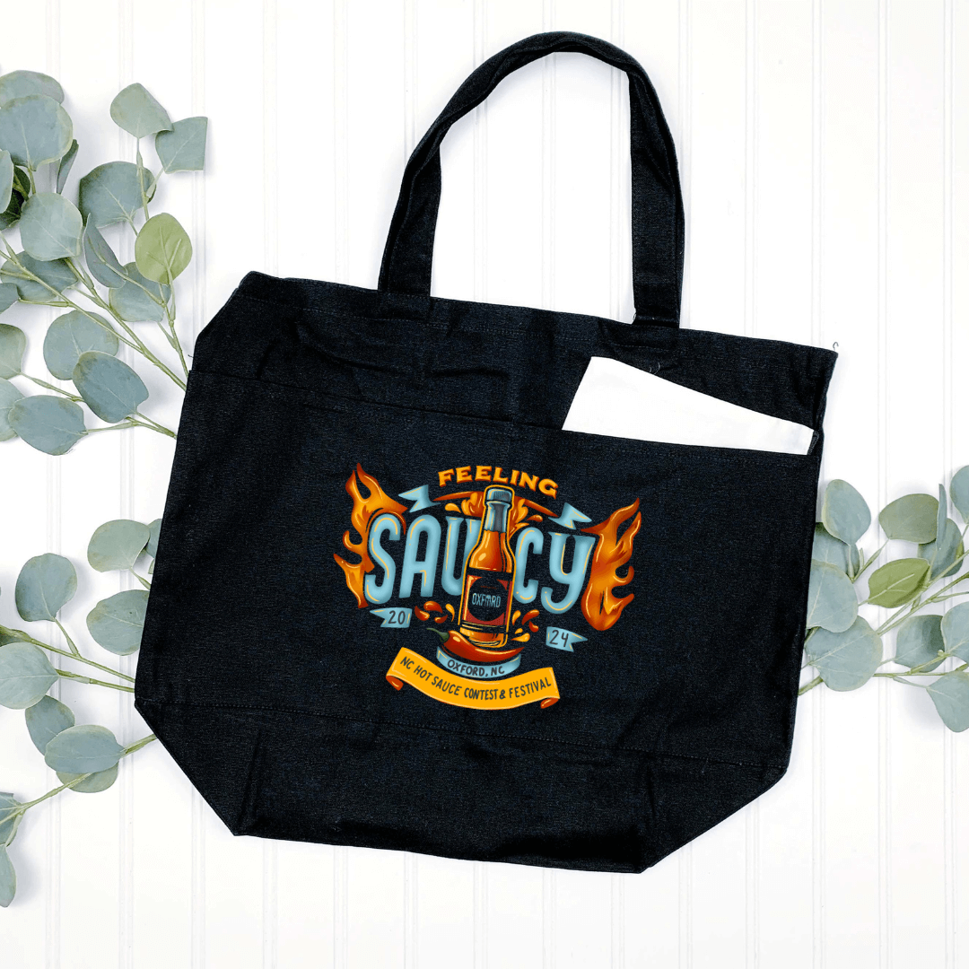 Black NC Hot Sauce Contest & Festival Tote Bag with "Feeling Saucy" design, surrounded by greenery on a white background.