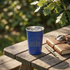 A blue insulated travel tumbler with a stainless steel rim sits on a wooden picnic table outdoors. The tumbler has placeholder text reading &