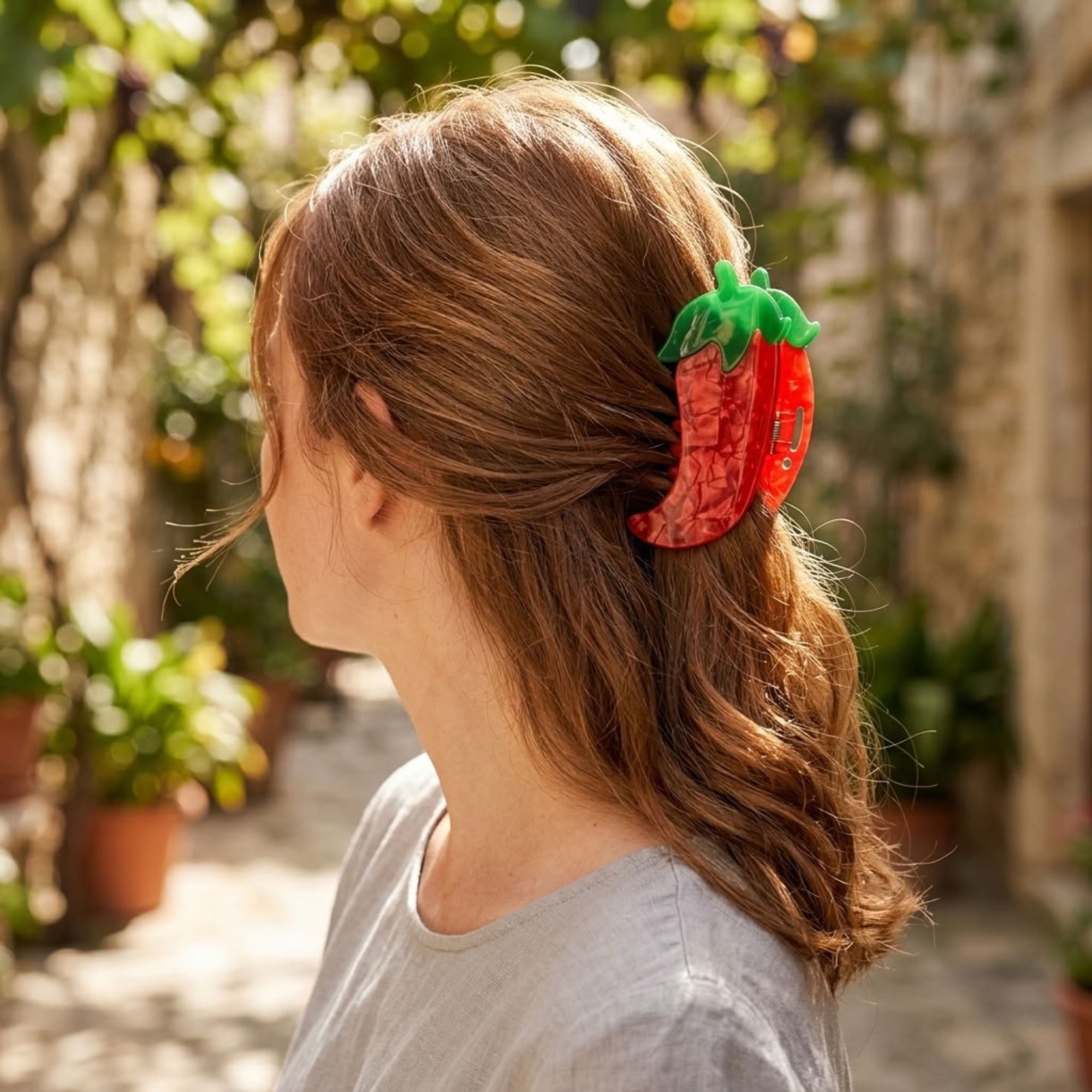 A woman with light brown hair is shown from the side in an outdoor garden setting, wearing a red chili pepper-shaped hair claw clip.