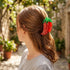 A woman with light brown hair is shown from the side in an outdoor garden setting, wearing a red chili pepper-shaped hair claw clip.