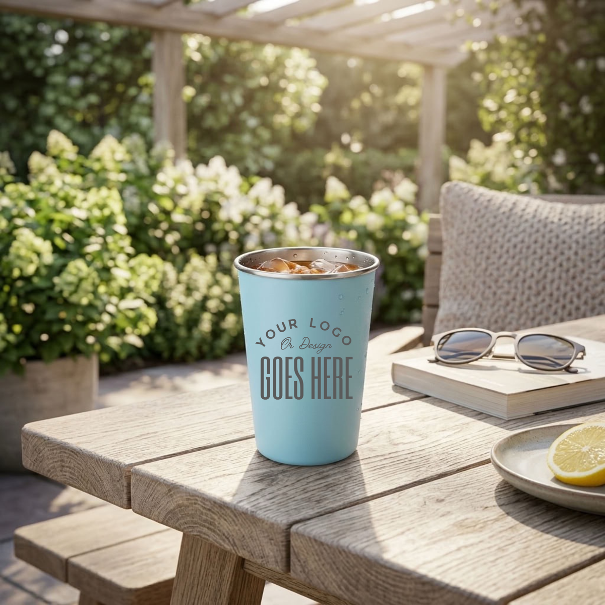 A light blue insulated travel mug with customizable text sits on a wooden outdoor table, surrounded by a book with sunglasses, a lemon slice, and a patio garden background.