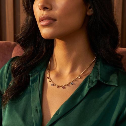 A close-up lifestyle shot of a woman wearing a delicate gold necklace with small dangling charms, paired with a dark green satin shirt.