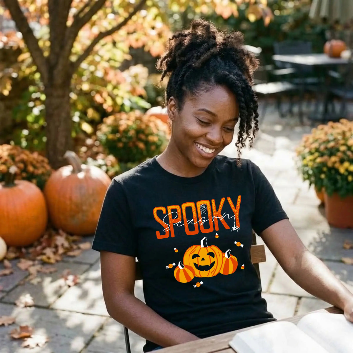 Smiling woman in a black "Spooky Season" Halloween graphic tee reading at a patio table surrounded by pumpkins