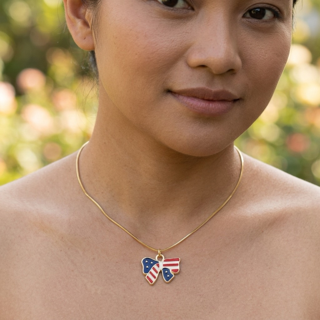 A close-up of a woman wearing a delicate gold chain necklace with a bow-shaped pendant decorated with the American flag pattern, set against a soft, blurred outdoor background.