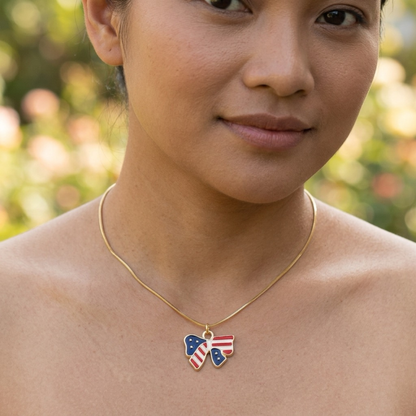 A close-up of a woman wearing a delicate gold chain necklace with a bow-shaped pendant decorated with the American flag pattern, set against a soft, blurred outdoor background.