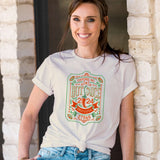 Woman wearing NC Hot Sauce Contest & Festival T-shirt featuring a vintage label design, standing by a stone wall.