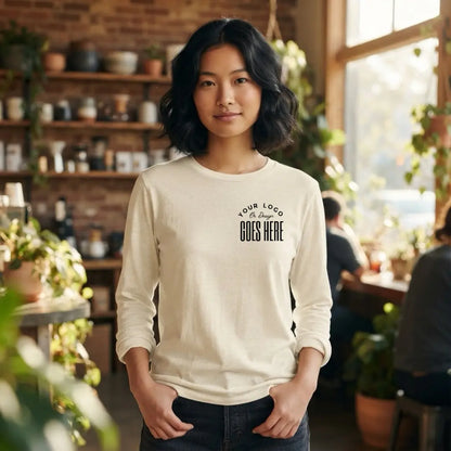 Cream custom long sleeve t-shirt with a black "Your Logo or Design Goes Here" left chest placeholder, worn in a plant-filled cafe