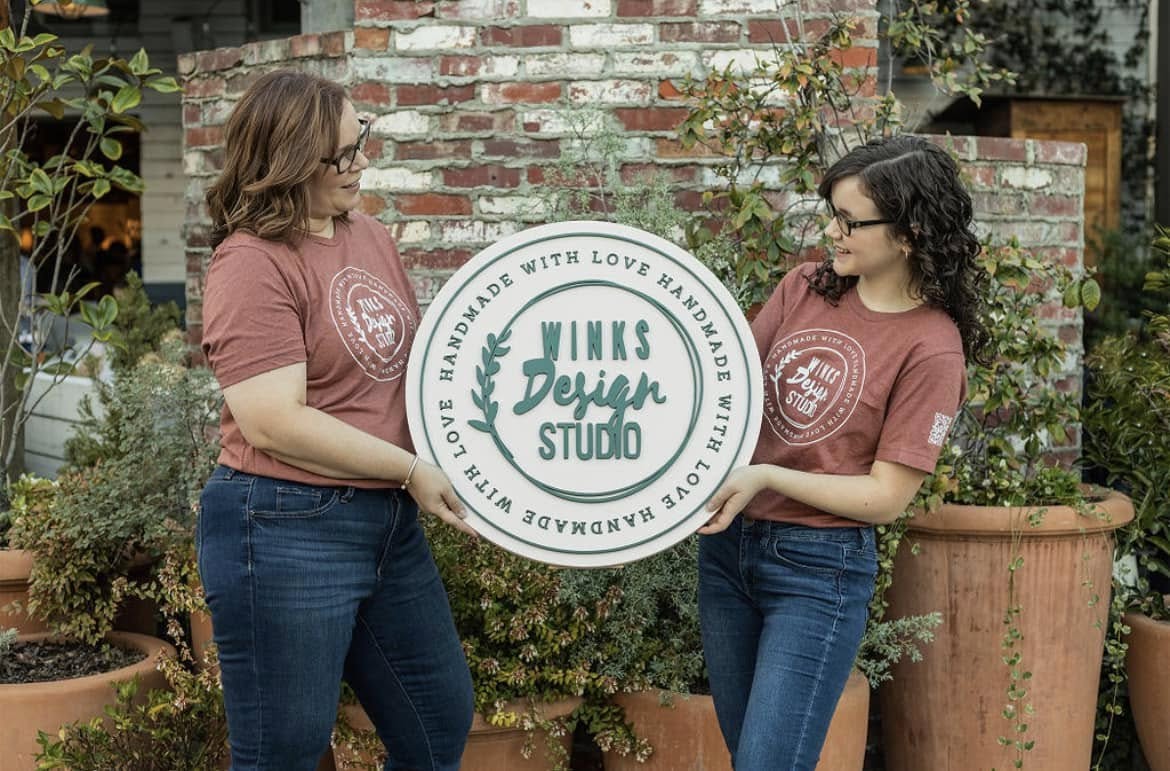 Two women holding a 'Winks Design Studio' sign in front of a brick wall.
