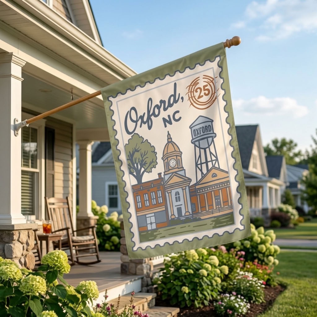 A decorative garden flag hanging from a wooden pole on a house porch. The flag has a vintage stamp-like border and an illustration of Oxford, NC buildings and trees, with the text &