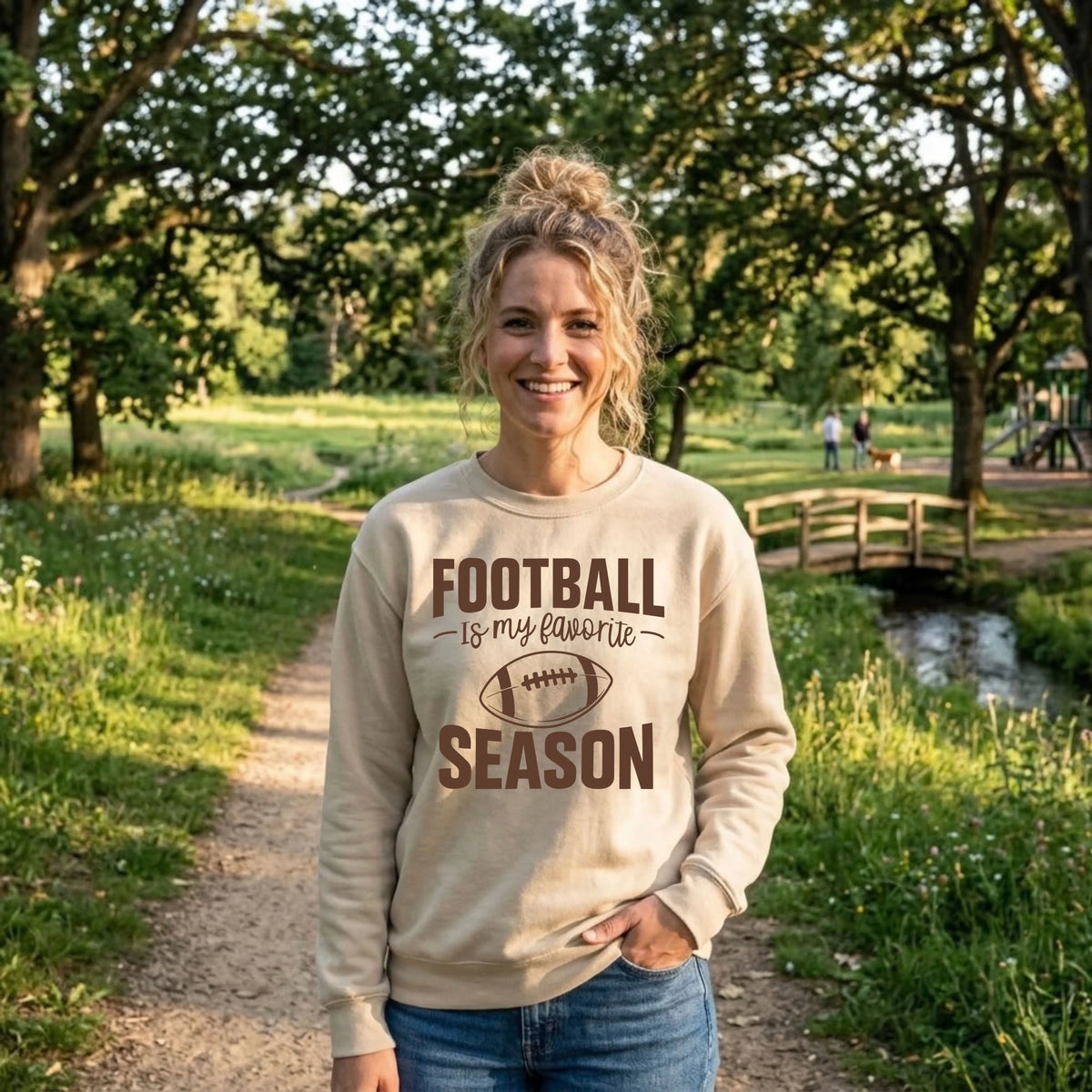 Smiling woman in a sand "Football Is My Favorite Season" graphic crewneck sweatshirt on a sunny park trail