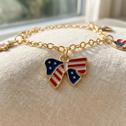 A gold chain bracelet with a bow-shaped American flag charm, resting on a beige textured fabric surface near a window.