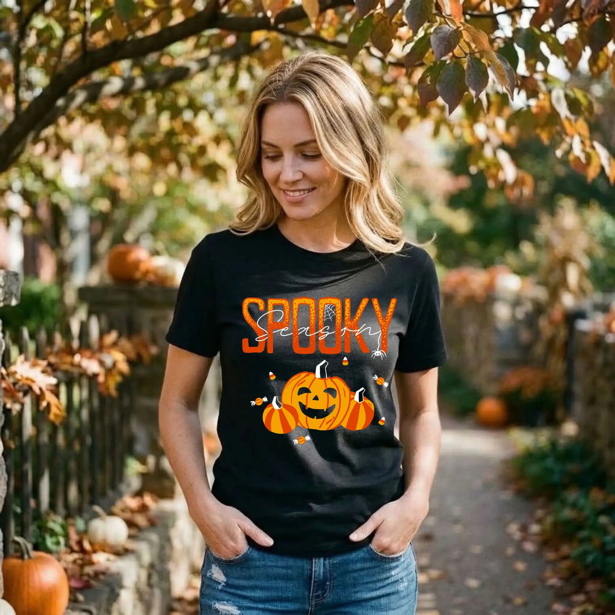 Woman in a black "Spooky Season" Halloween graphic tee and jeans on an autumn lane lined with pumpkins