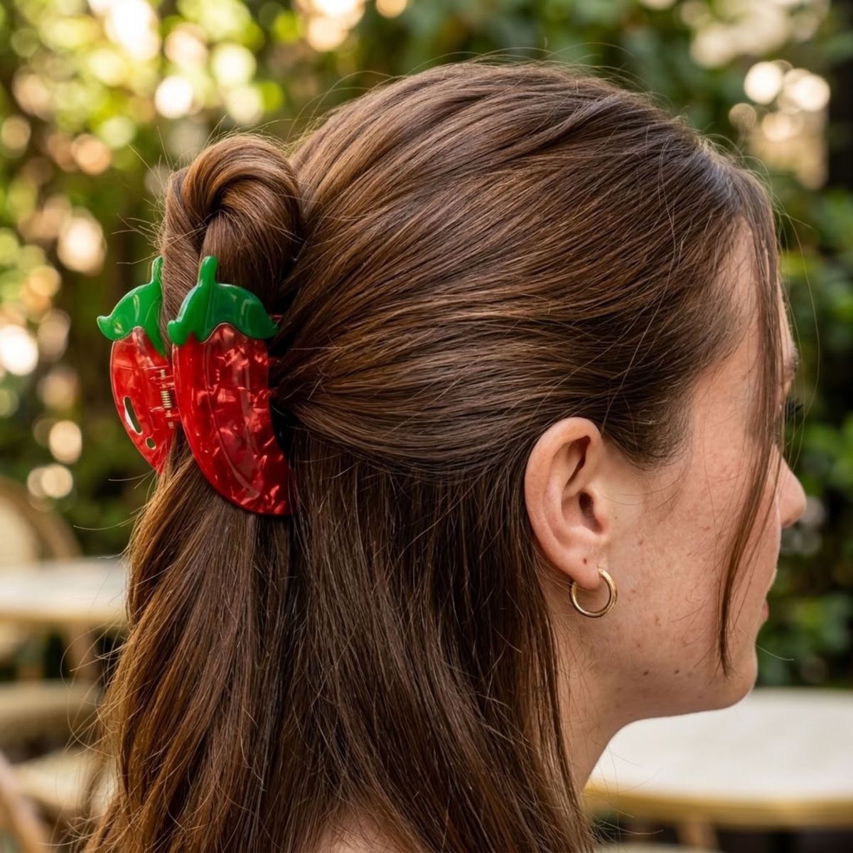 A woman with brown hair in a half-up bun wearing a red strawberry-shaped hair claw clip with green leafy tops, photographed outdoors with a blurred green background.
