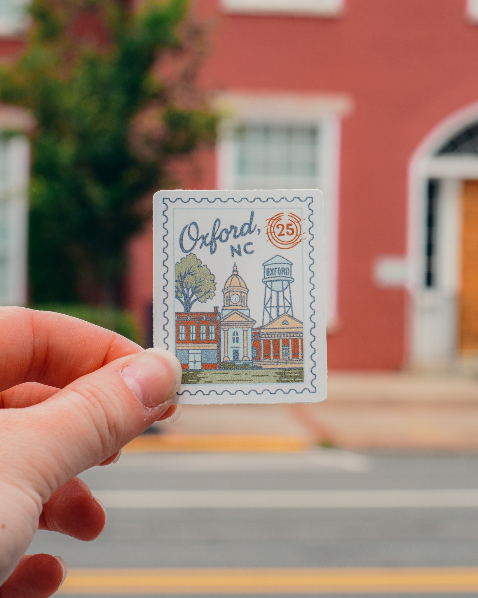 Hand holding a postcard with Oxford, NC design in front of a red building.