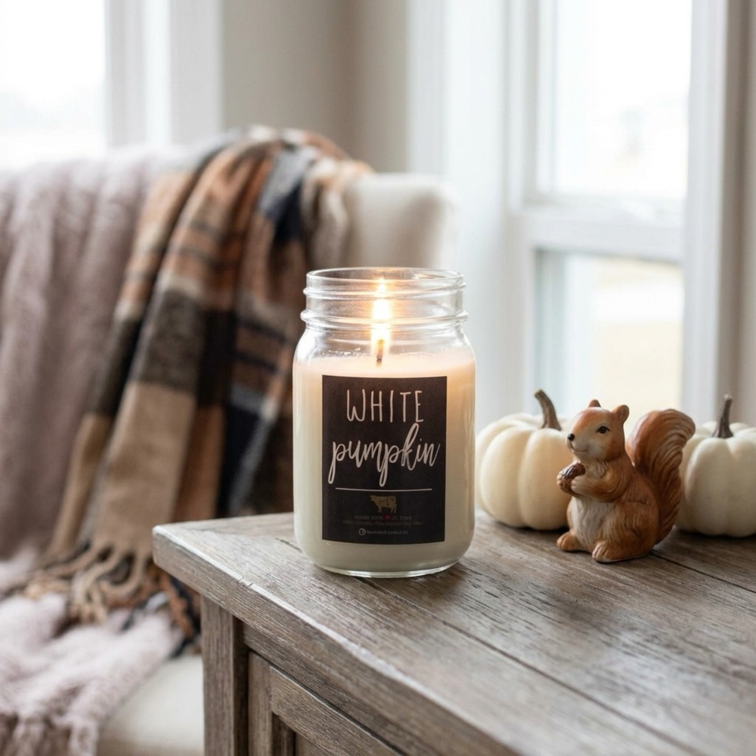 A lit white candle in a clear glass mason jar sits on a rustic wooden table in a cozy indoor setting. The front label reads “WHITE pumpkin.