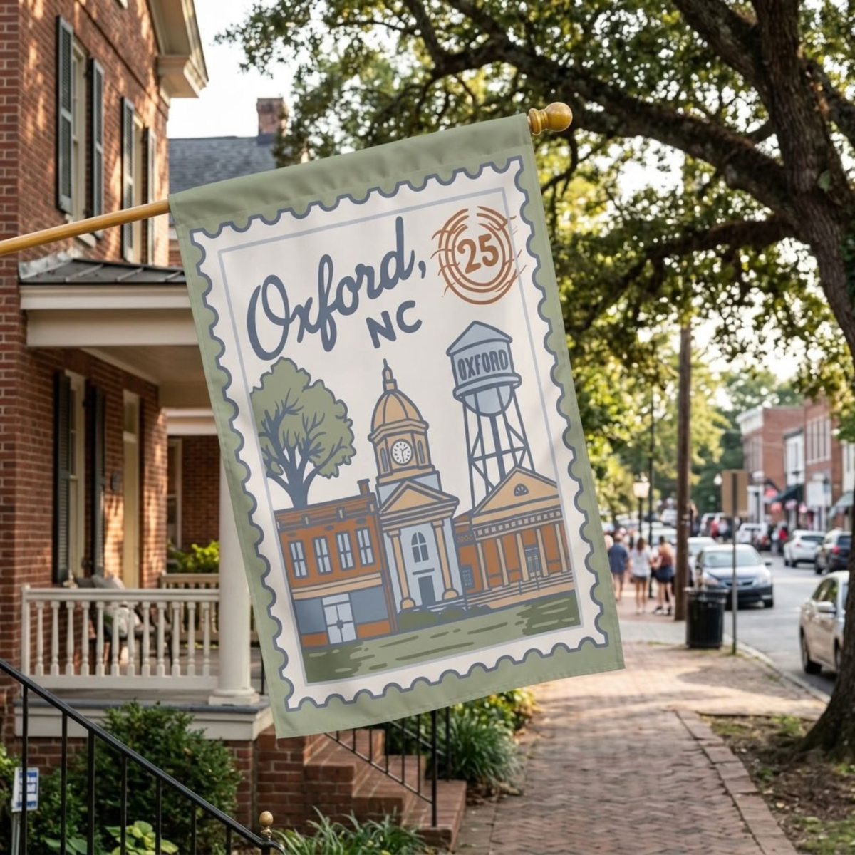 A decorative garden flag hanging from a wooden pole on a brick house porch, featuring an illustrated Oxford, NC stamp-style design with buildings, trees, and a water tower. The scene is set outdoors on a sidewalk with trees and street activity in the background.