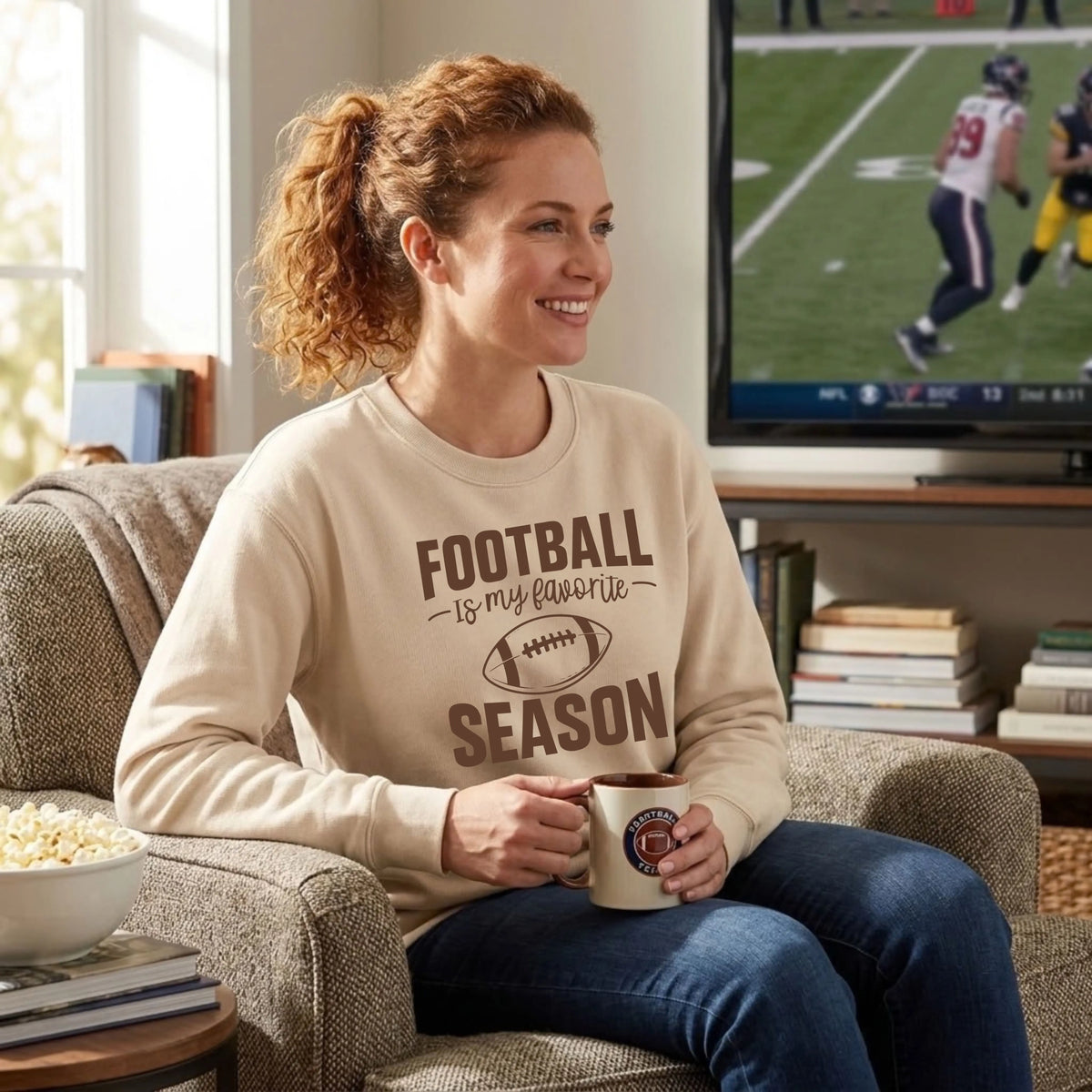 Woman in a sand "Football Is My Favorite Season" graphic crewneck sweatshirt watching a game at home