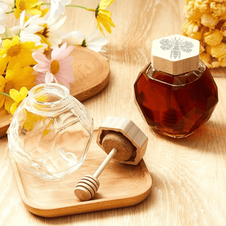 Engraved hexagon glass honey dispenser with dipper and wooden lid, surrounded by flowers, on a wooden tray.