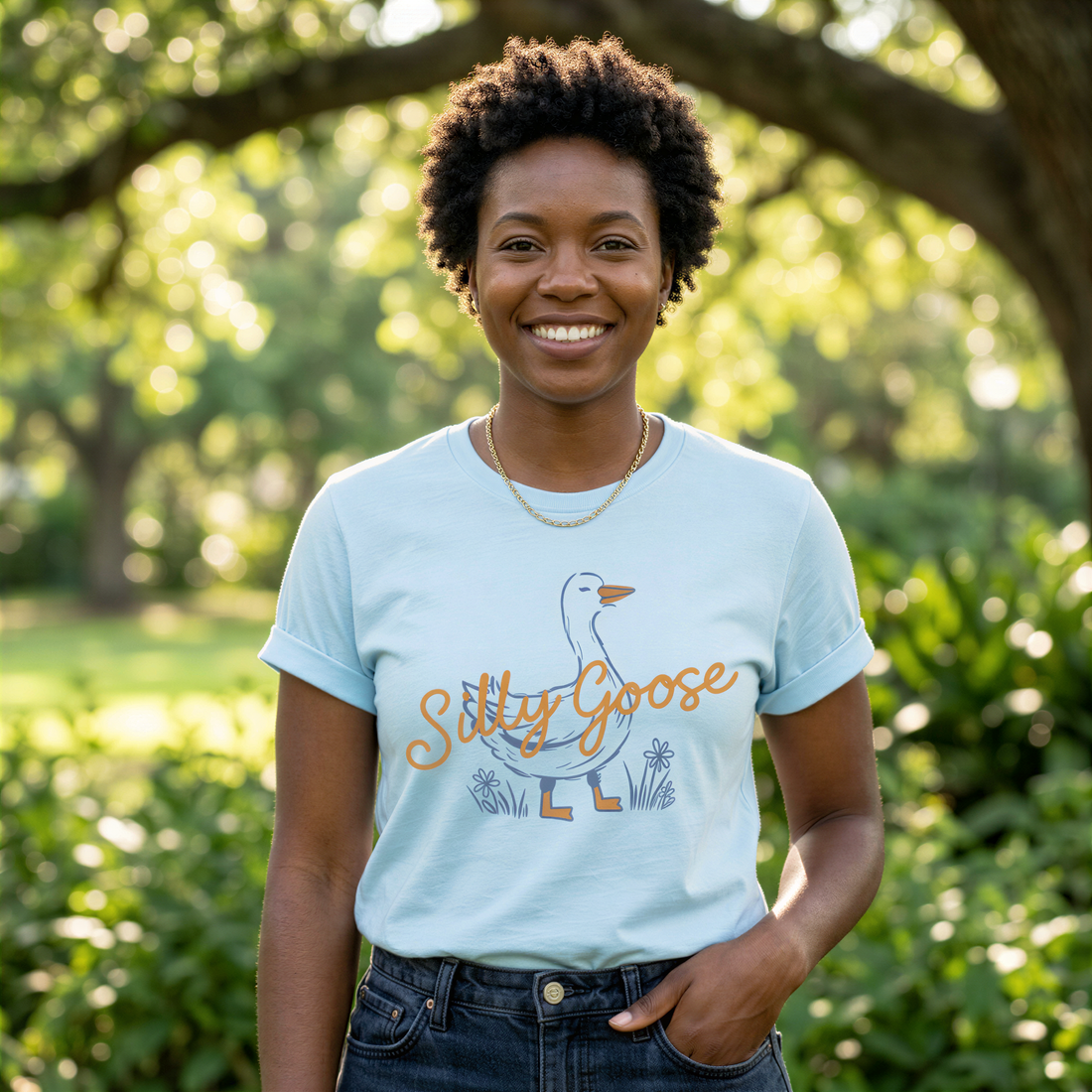 A smiling woman wearing a light blue t-shirt with a graphic of a goose and the text "Silly Goose" stands in a park surrounded by greenery.