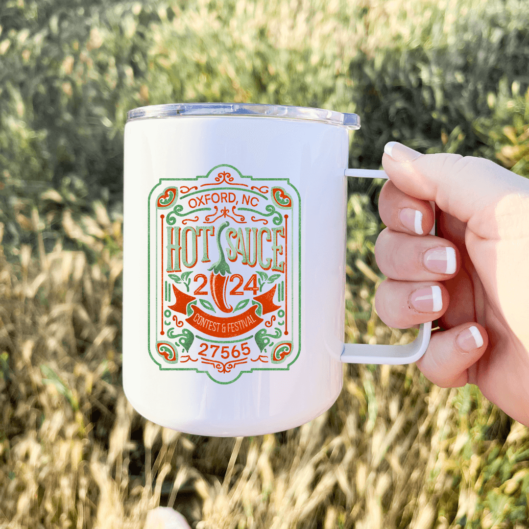 Person holding white 13oz mug with 2024 NC Hot Sauce Contest & Festival print in grassy field.