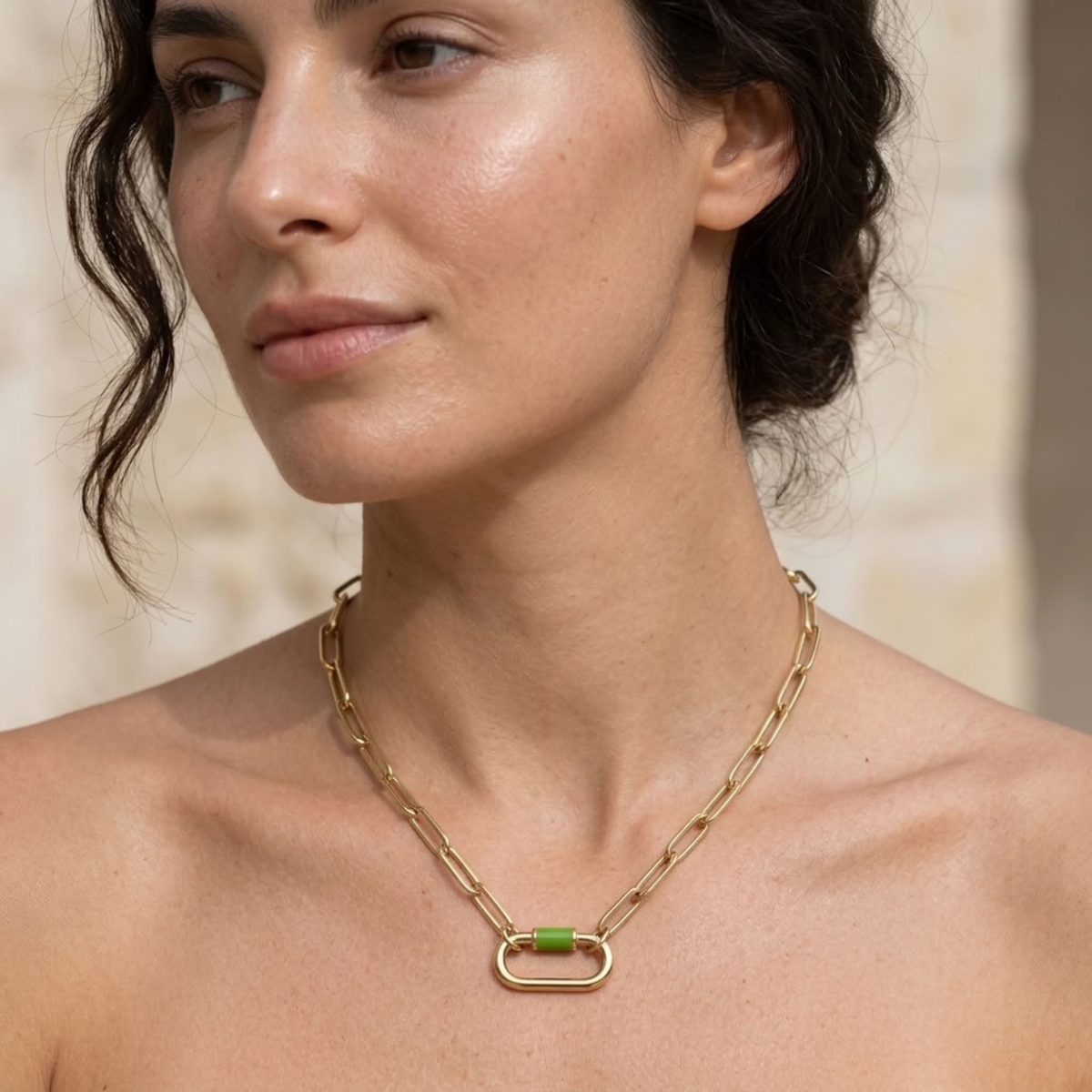 A close-up of a woman wearing a gold chain necklace with a capsule-shaped pendant featuring a small green accent, set against a softly blurred beige background.