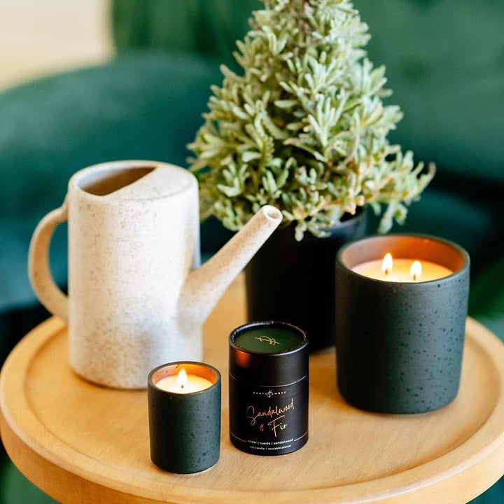 Decorative setting with candles, a watering can, and a small tree on a wooden table.