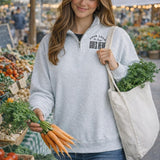 A woman is holding a bunch of carrots and a bag of greens, possibly at a farmer's market, with a sign on her back that says "Your logo goes here".