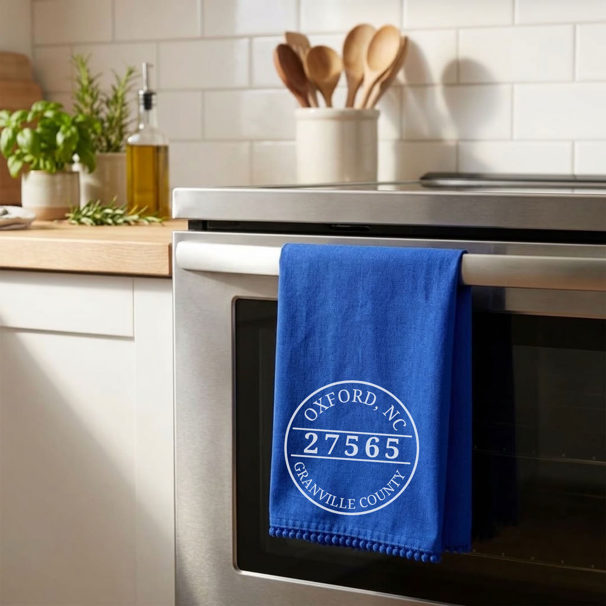 A blue kitchen towel with a white circular Oxford, New York address print hangs from the handle of a stainless steel oven in a bright, modern kitchen.