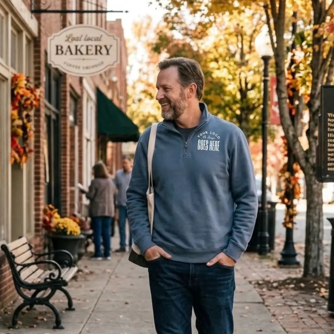 Slate blue custom quarter zip sweatshirt with a white "Your Logo or Design Goes Here" left chest placeholder on a downtown street