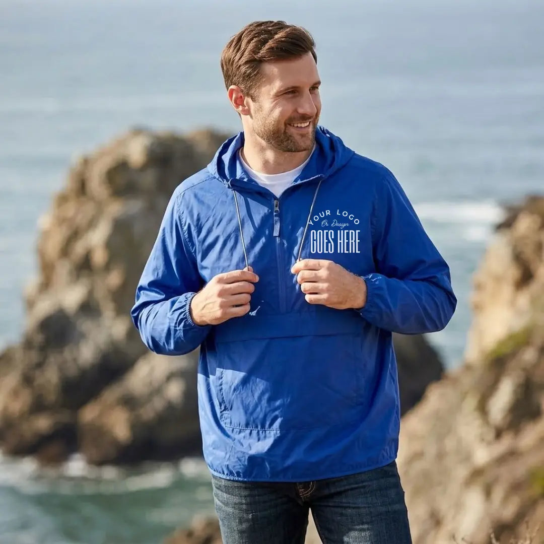 Man in a royal blue custom quarter zip windbreaker with a white "Your Logo or Design Goes Here" left chest placeholder on a seaside cliff