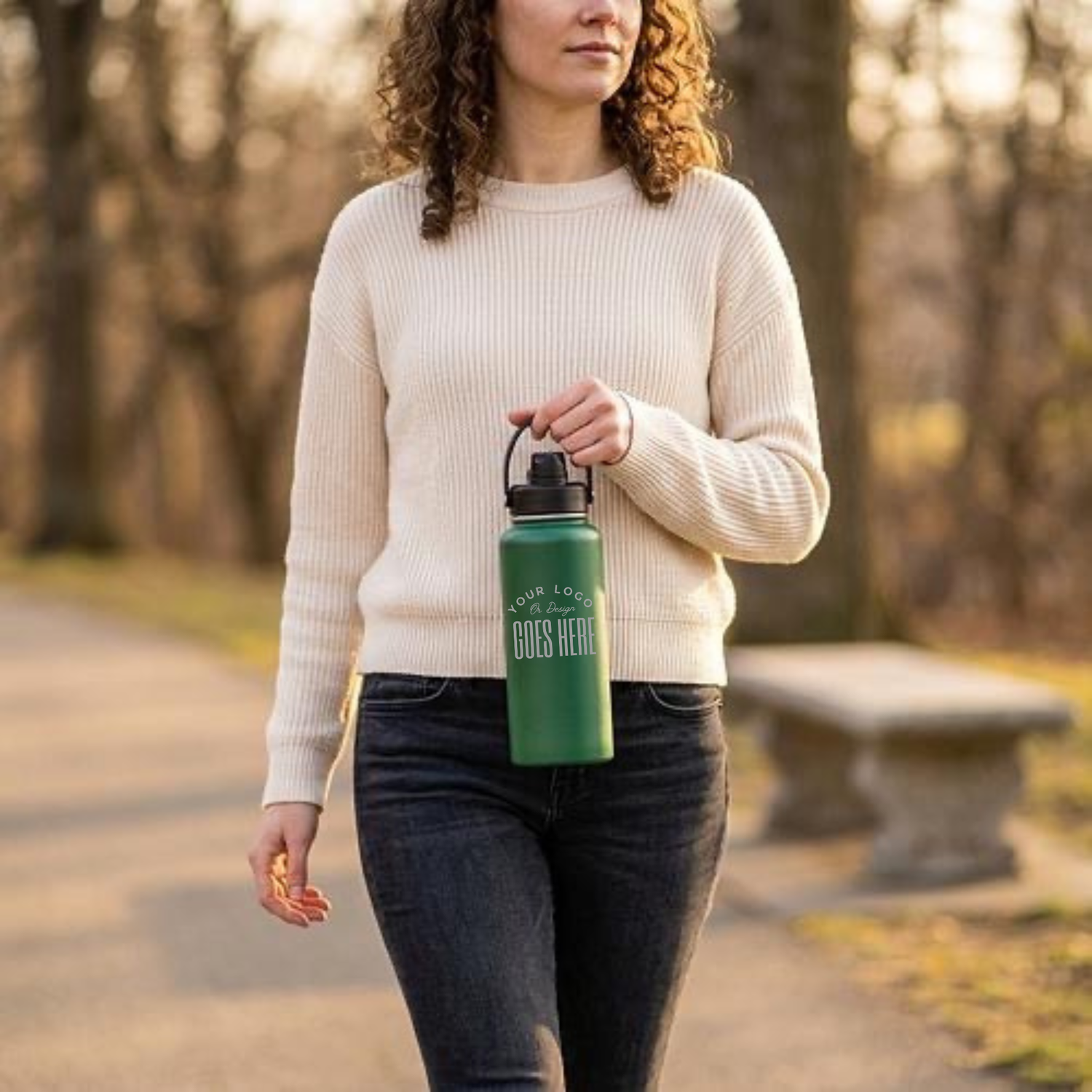 A woman walking outdoors in a park wearing a cream ribbed sweater and dark jeans, holding a green reusable water bottle with the words &